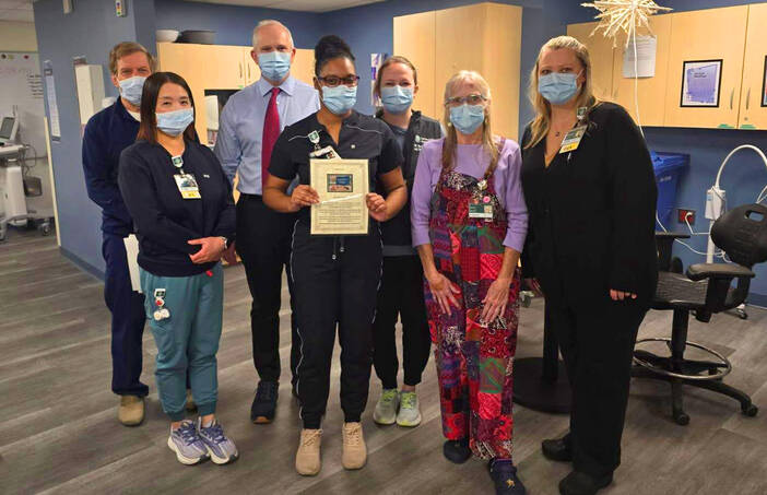 Seven healthcare providers in scrubs and masks stand in a room; one in the center holds a plaque.